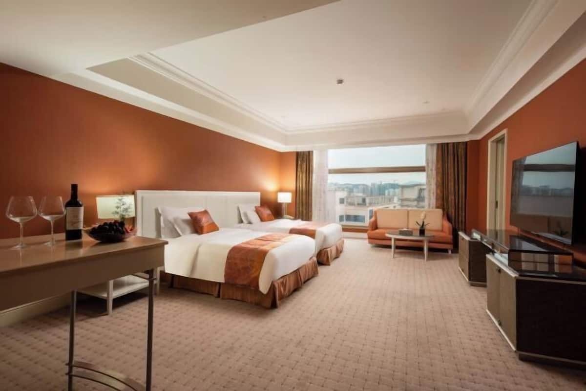 A hotel room with two beds, a desk, a television, and a view of the city.