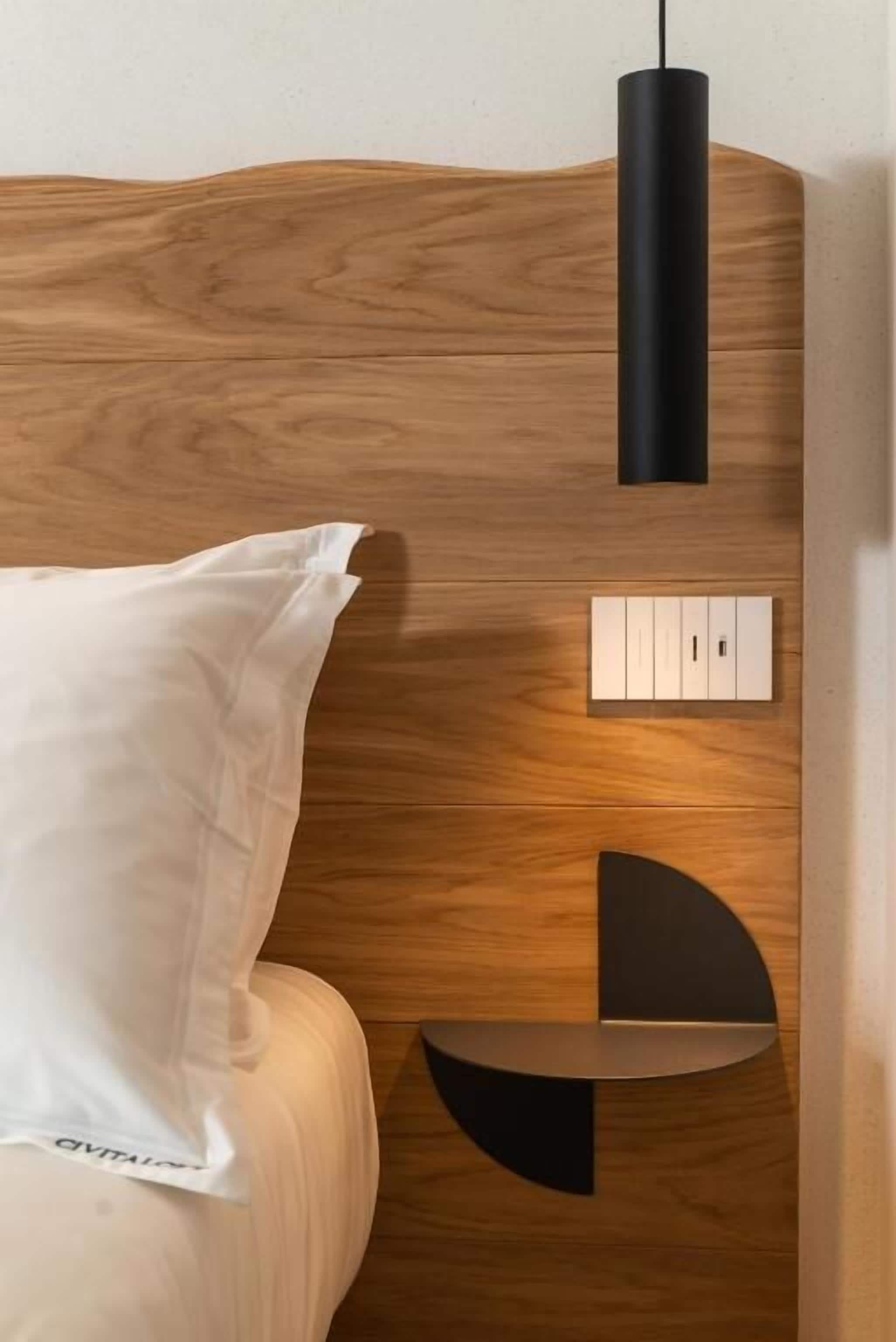 Una cama con cabecera de madera, almohada blanca, lámpara colgante negra y panel de control.