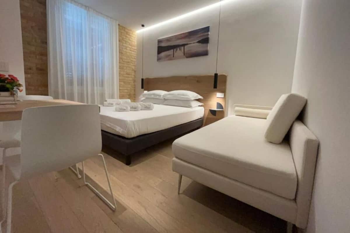 A modern hotel room with a wooden accent wall, a large bed, a bedside table with a lamp, and a white chair.