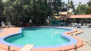 Una piscina con borde curvo, rodeada de sillas reclinables y mesas, con árboles y edificios al fondo.