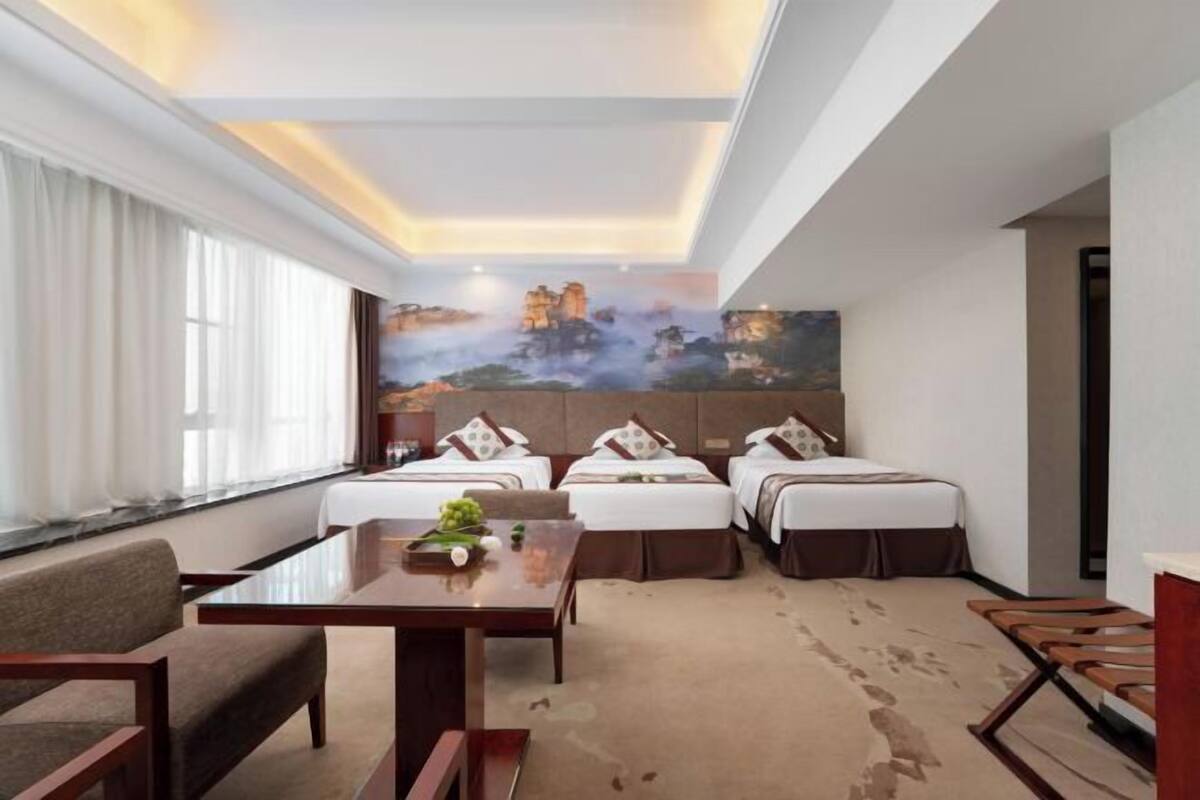 A hotel room with a large bed, a smaller bed, a desk, and a chair. There is a painting on the wall and a window with curtains.