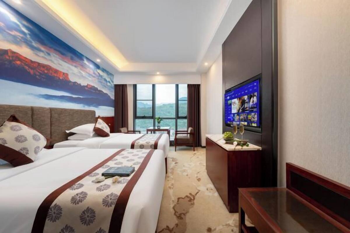 A hotel room with two beds, a desk with a TV, and a large window with a mountain view.