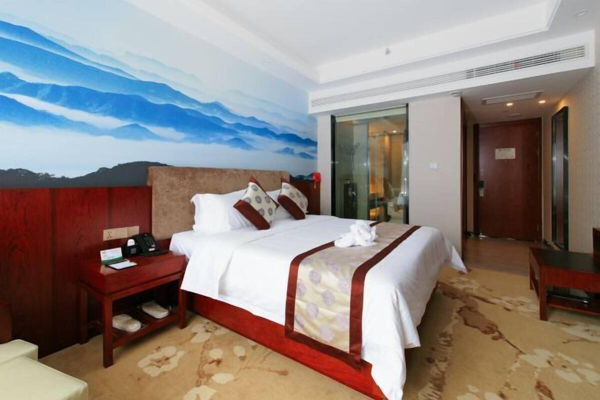 A hotel room with a large bed, a nightstand, a wooden headboard, a carpeted floor, and a wall mural.
