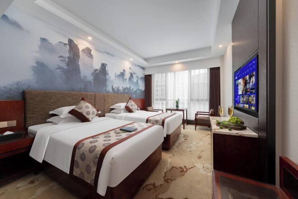 A hotel room with two beds, a large wall mural, a flat-screen TV, and a small table with a wine glass and grapes.