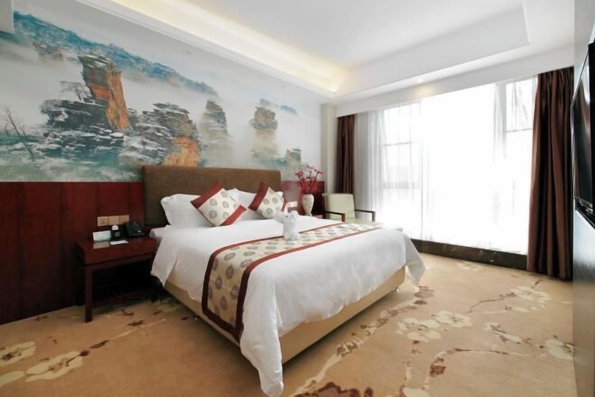 Una habitación de hotel con una cama grande, una mesita de noche, un cuadro en la pared y una ventana con cortinas.