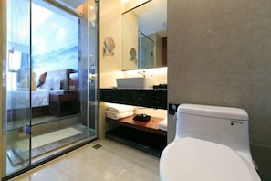 Un baño de hotel moderno con cabina de ducha de vidrio, inodoro blanco, lavabo con espejo y vistas a una cama a través del vidrio.