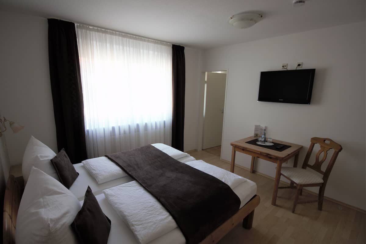 A hotel room with a bed, a TV mounted on the wall, a small table with a chair, and a window with curtains.