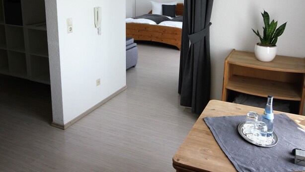 Moderni hotellihuone, jossa on puinen ruokapöytä, tuoli, pieni ruukkukasvi ja tummalla verholla varustettu sänky.