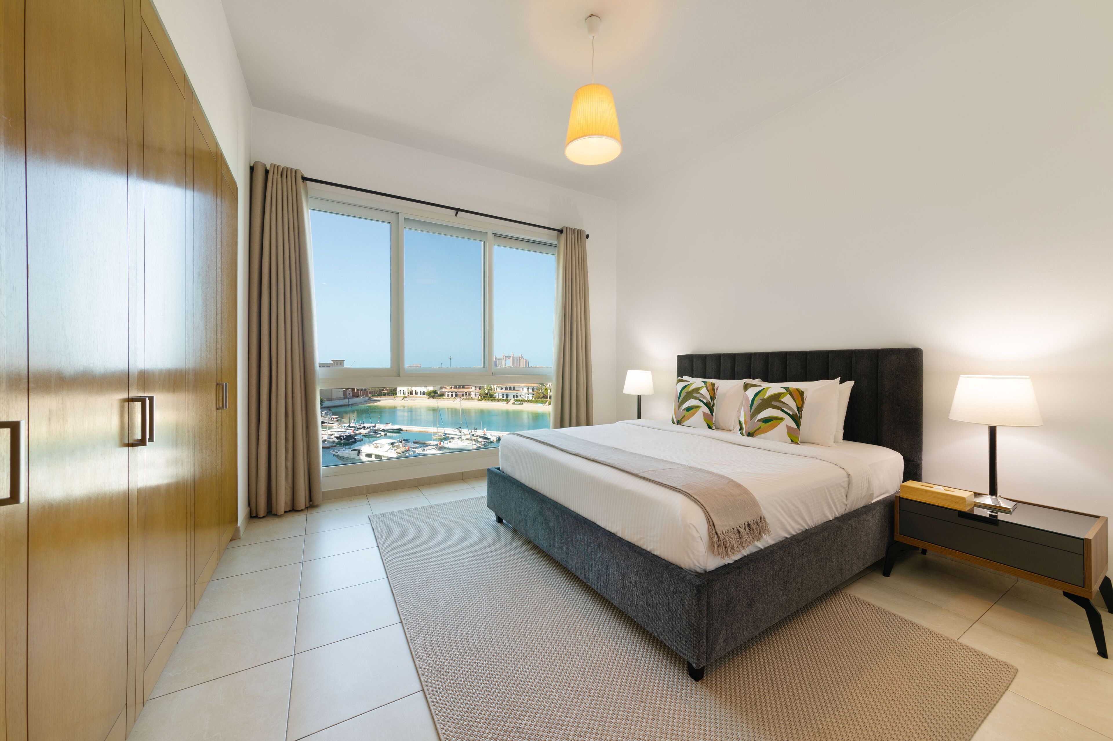 Une chambre moderne avec un grand lit, une table de chevet, une lampe et une vue sur une piscine et des immeubles.