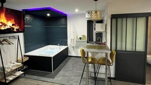 A modern room with a large flat-screen TV, a bathtub, a dining area with two chairs, and a small kitchen area.