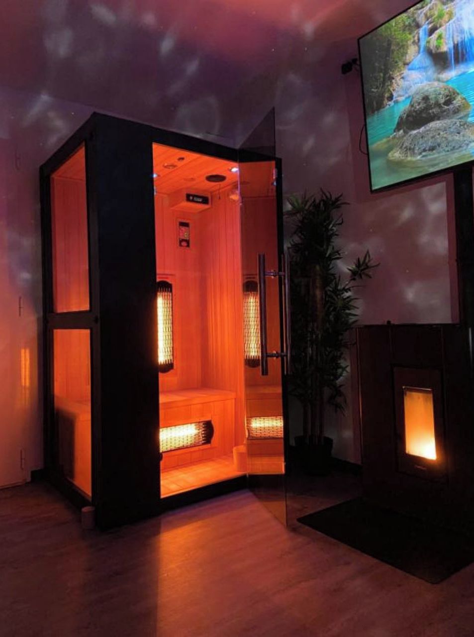 Un sauna con suelo de madera, puerta de vidrio y televisor en la pared mostrando una escena de cascada.