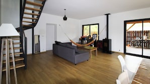 A spacious living area with a wooden staircase, a grey sofa, a hammock, and a large window with a view of bicycles outside. - Pretty wooden house 5 minutes from the ocean (Labenne)
