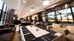 Un hall d’hôtel spacieux, avec une longue table à manger dressée pour le repas, des sièges confortables et de grandes fenêtres offrant une vue panoramique.