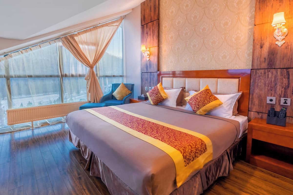 A modern hotel room with a large bed, a wooden headboard, a view of the outdoors through large windows, and a small desk with a chair.