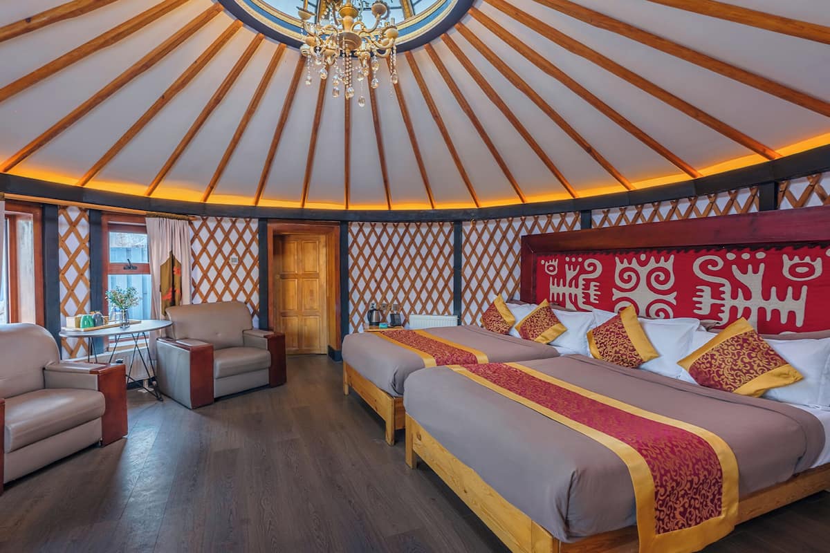 A room with a high ceiling, wooden beams, a chandelier, a bed with patterned bedding, a sofa, and a small table with a vase of flowers.