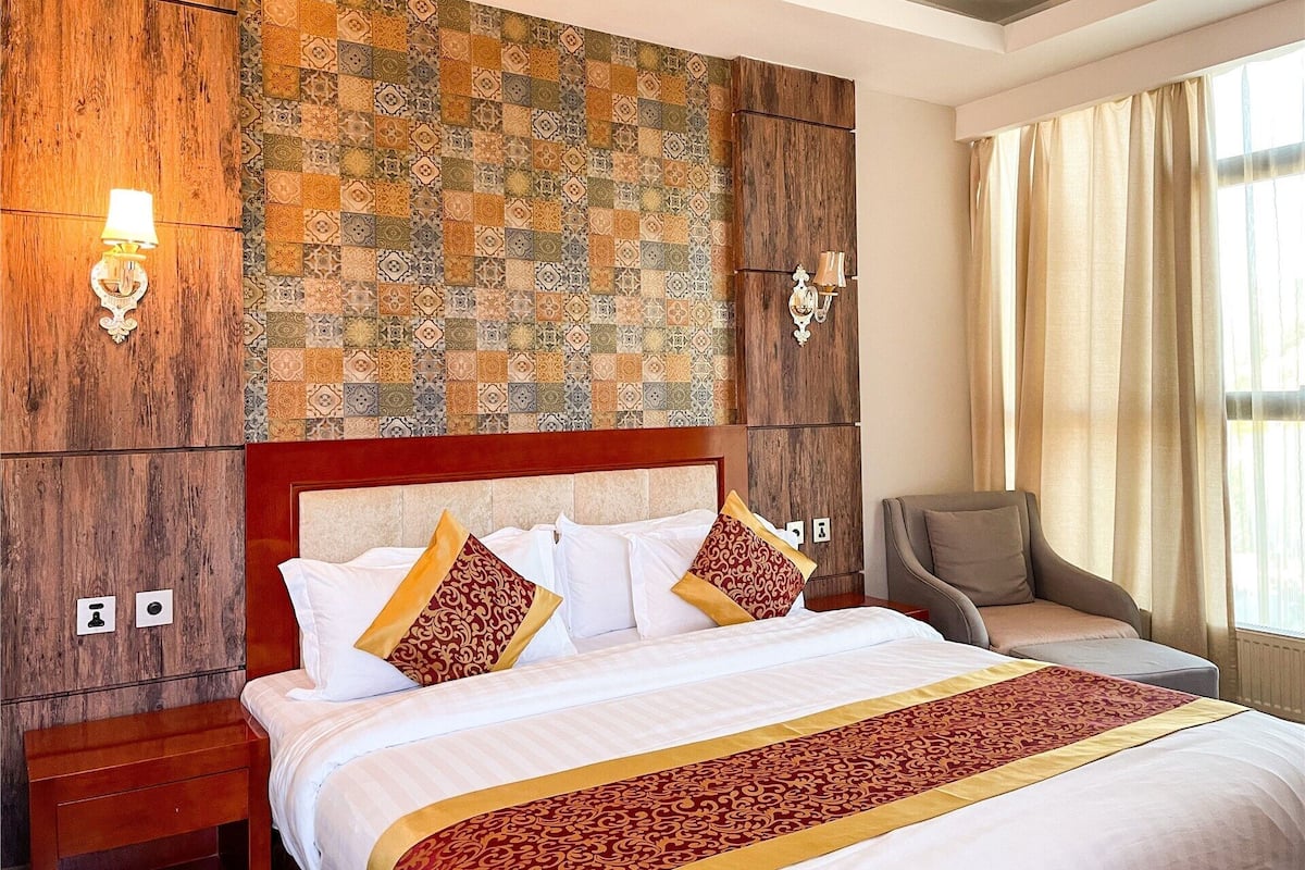 A hotel room with a large bed, a wooden headboard, patterned wall tiles, two bedside lamps, a chair, and a window with curtains.