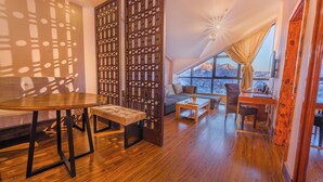 Un salon chaleureux avec une table en bois, un banc et un canapé, un paravent décoratif et une grande fenêtre donnant sur une vue imprenable sur les montagnes.