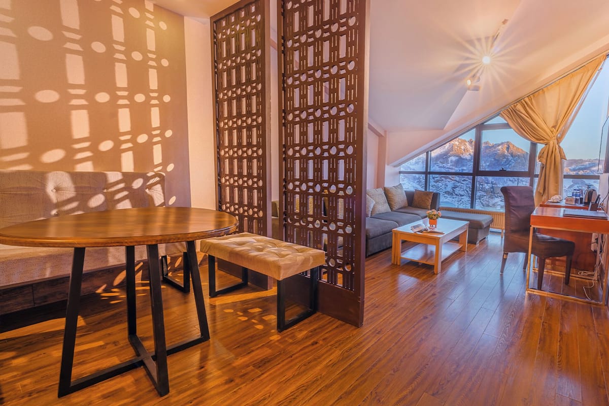 A cozy living area with a wooden table, bench, and sofa, a decorative screen, and a large window offering a mountain view.