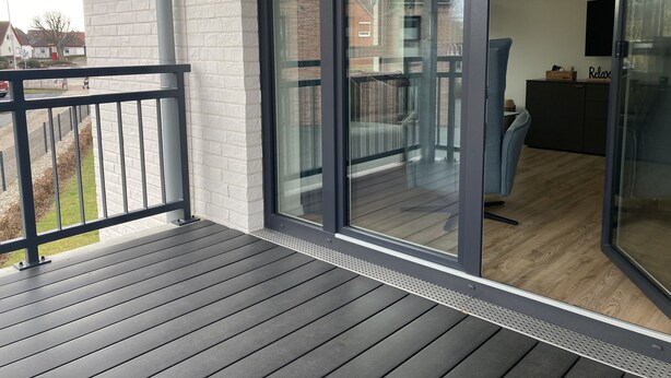 Ein Balkon mit Holzboden, eine Glastür zu einem Zimmer mit einem dunkelgrauen Sofa und einem kleinen Tisch.