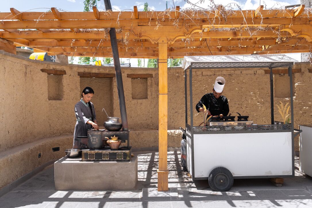 A traditional cooking setup with a woman preparing food and a man grilling under a wooden pergola.