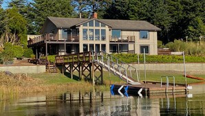 A two-story house with a dock extending into a lake, surrounded by dense forest. - Luxurious Getaway on the Lake with hot tub, pool table, boat dock, covered porch (Langlois)