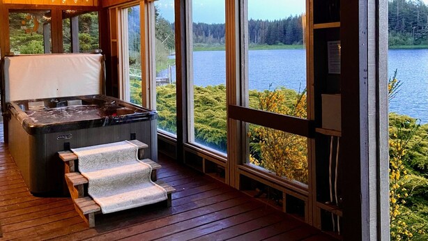 A hot tub with steps, wooden deck, and large windows overlooking a lake. - Luxurious Getaway on the Lake with hot tub, pool table, boat dock, covered porch (Langlois)