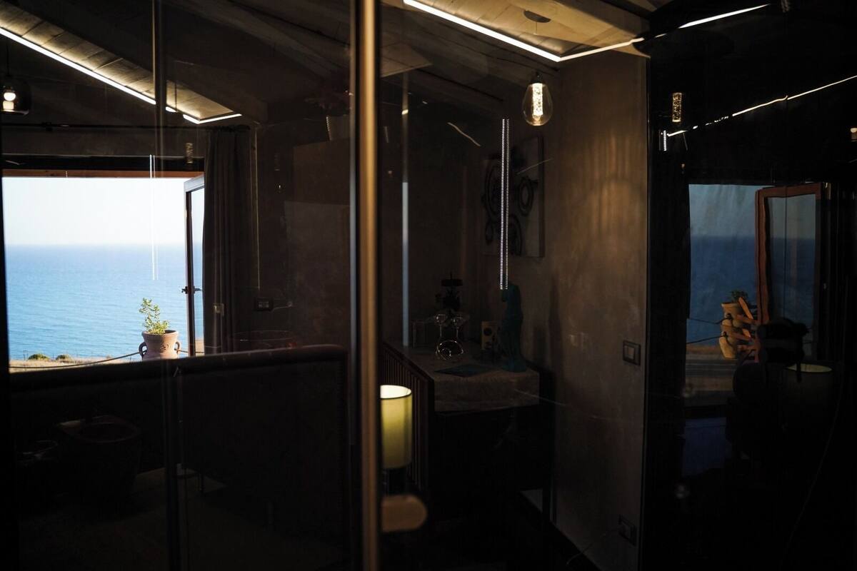 Una habitación tenuemente iluminada con vista al océano a través de una ventana, una pequeña mesa con una lámpara y una estatua.