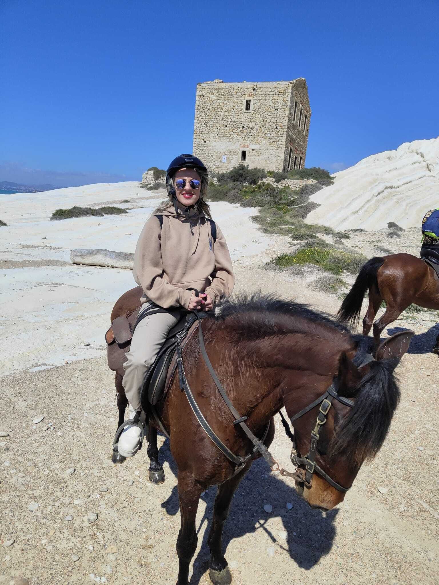 Eine Person reitet auf einem Pferd auf einem Sandweg, im Hintergrund ein Steinhaus.