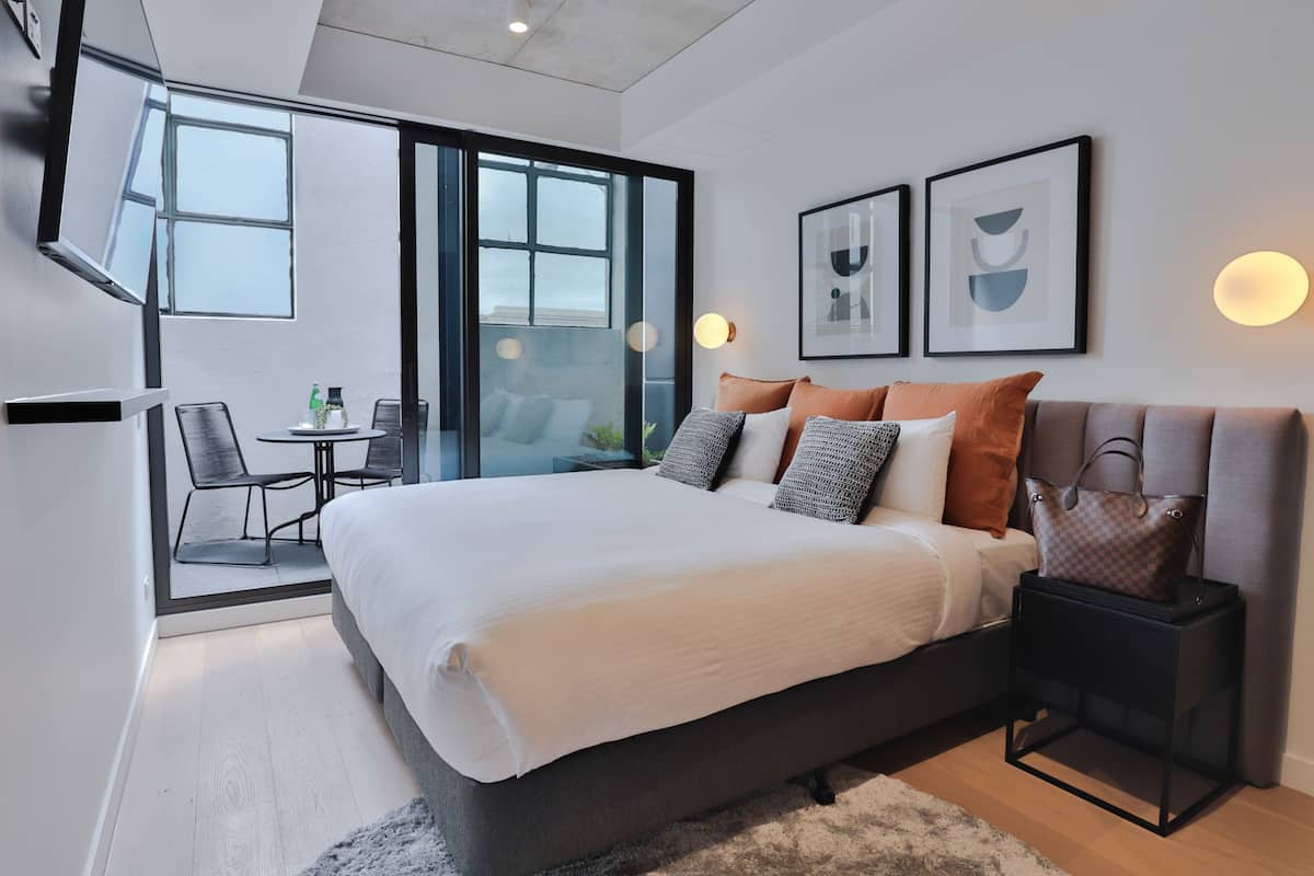 Un dormitorio moderno con una cama grande, un ventanal de vidrio que da a un balcón y una pequeña mesa con sillas.