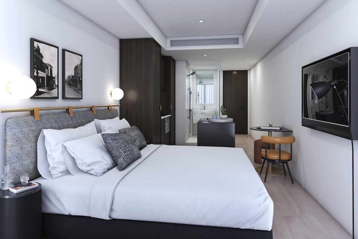 A modern hotel room with a large bed, bedside tables, a desk, a chair, a mirror, and framed pictures on the wall.