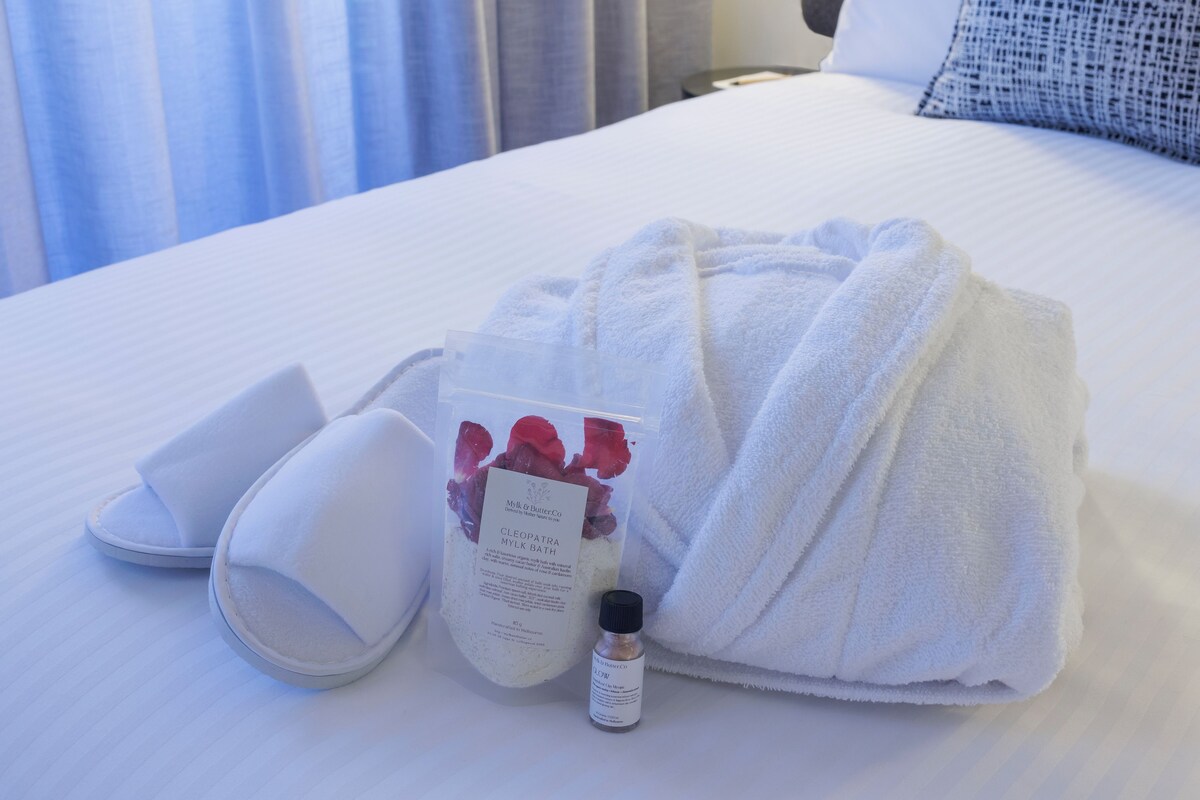 A neatly made bed with a white towel, slippers, a bottle, and a bag of bath products.