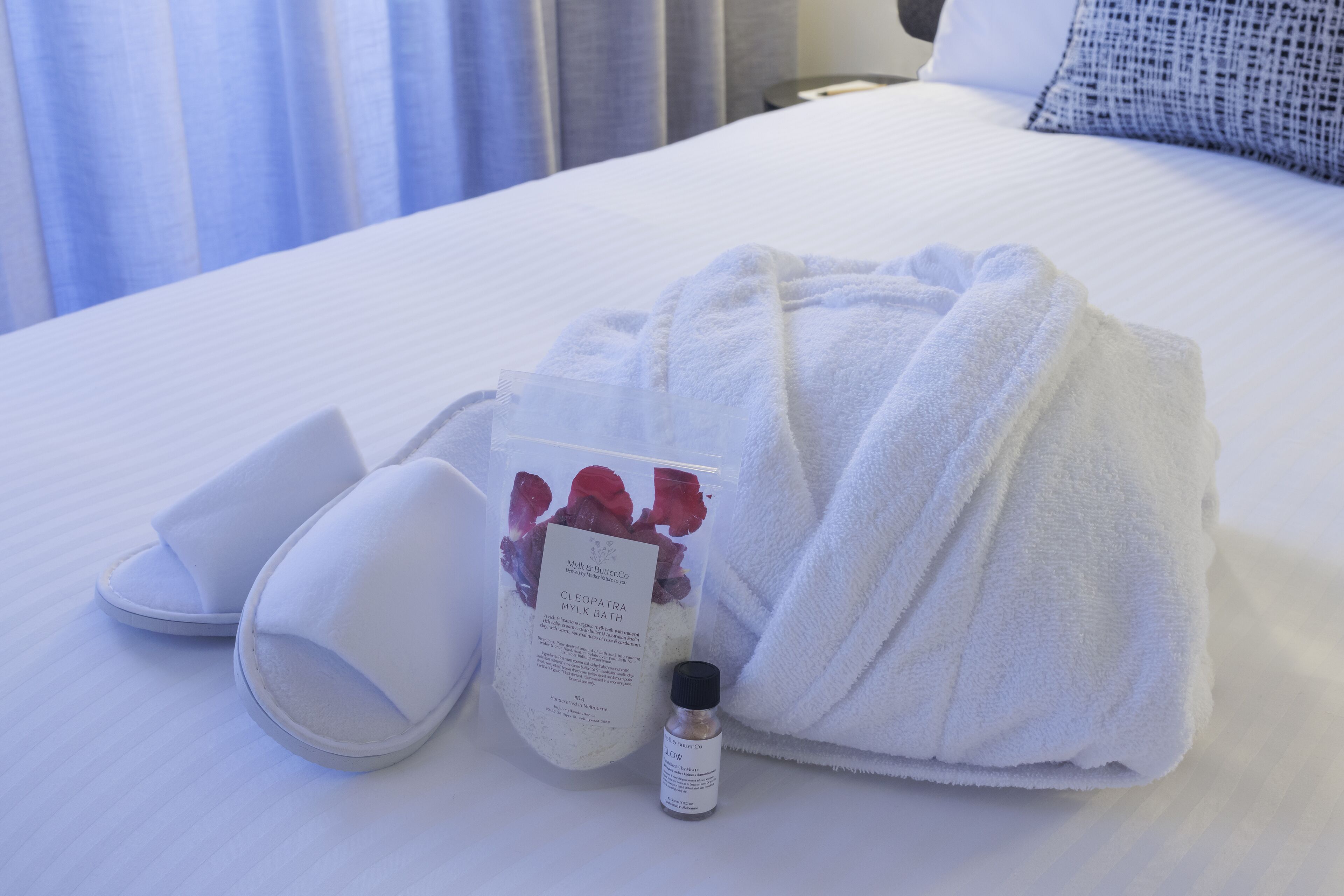 Une chambre d’hôtel avec un lit parfaitement fait, un peignoir blanc, des chaussons, une bouteille de produit de bain et un sac contenant des articles de toilette.
