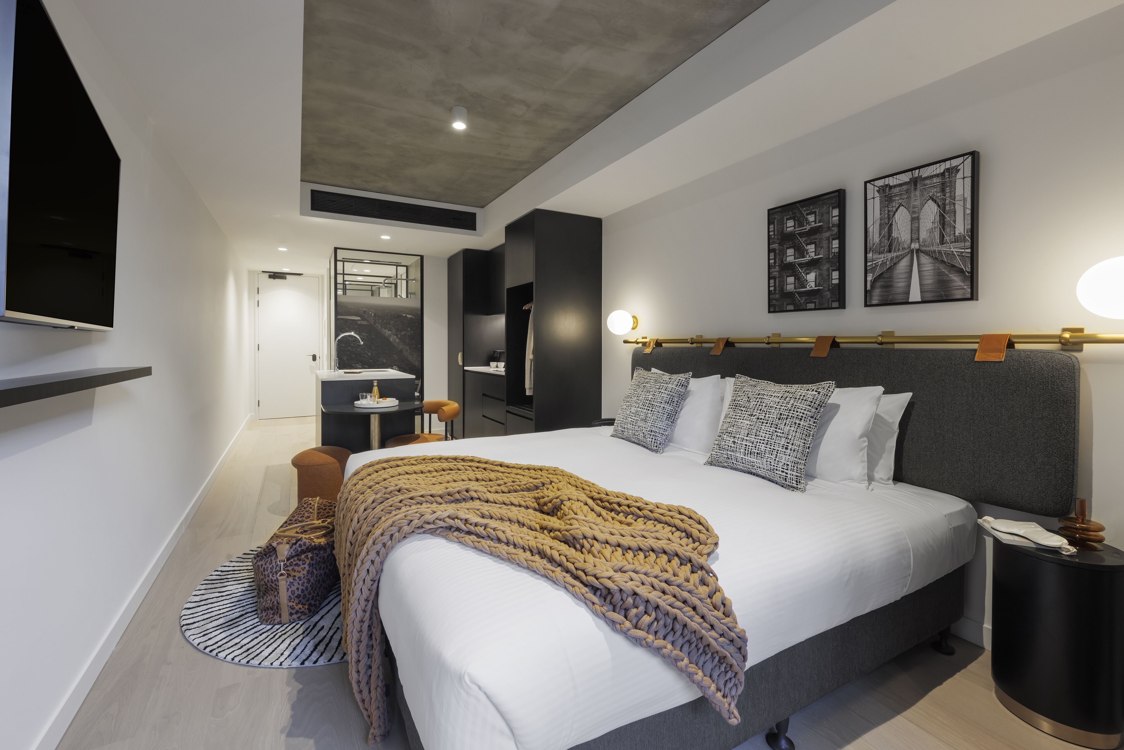 A modern bedroom with a large bed, a bedside table, a nightstand, a chair, and a bathroom visible in the background.