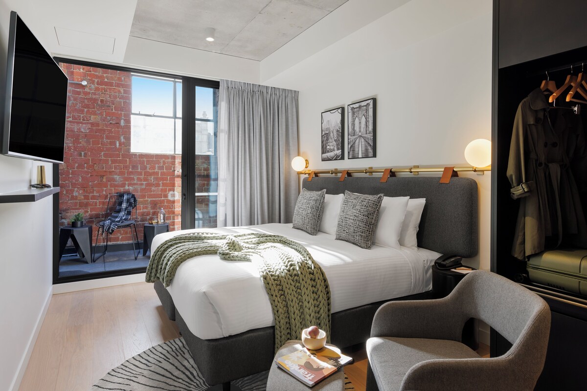 A modern hotel room with a large bed, a television, a seating area with a chair, and a view of the city through a large window.