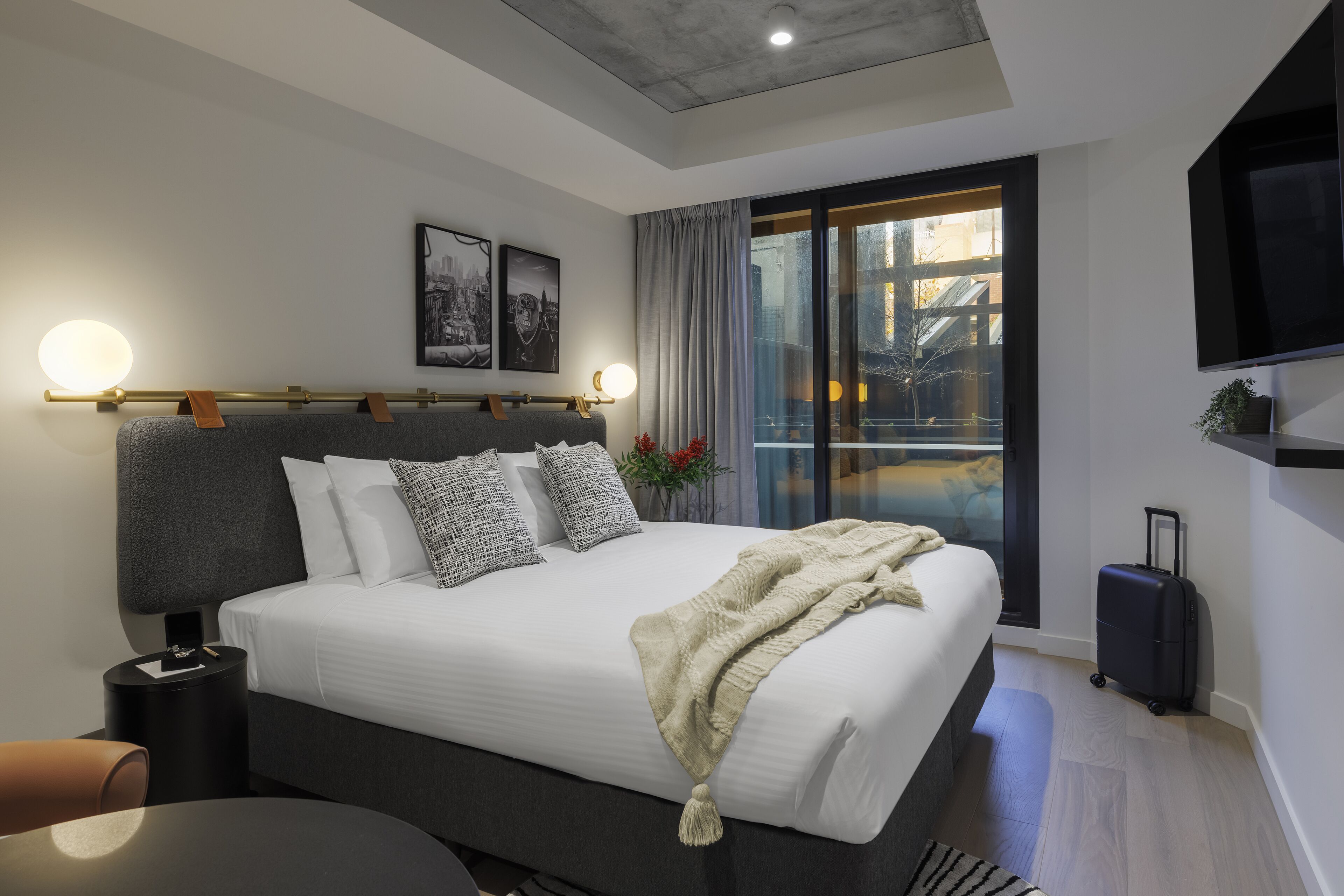 A modern hotel room with a large bed, a bedside table, a TV mounted on the wall, and a view of the city through the window.