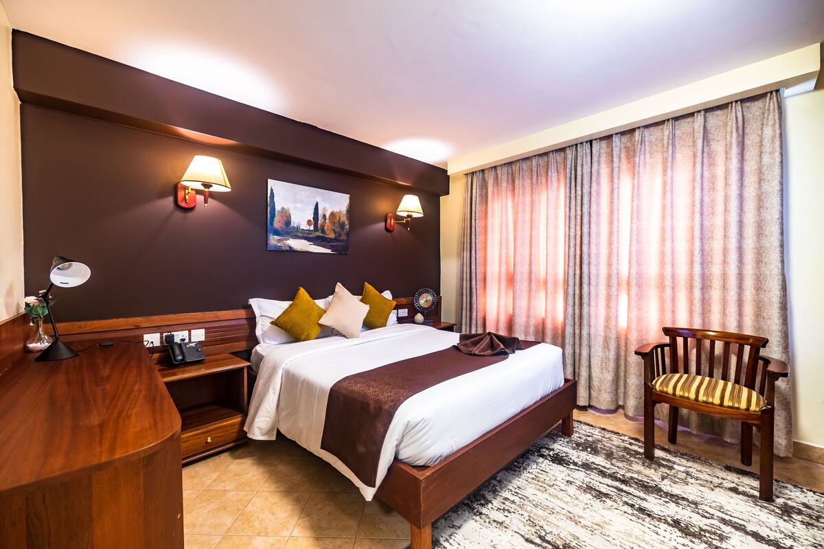 A hotel room with a large bed, bedside tables, a desk, and a chair. There are two wall-mounted lamps and a painting above the bed.
