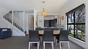 Una cocina moderna con una isla central, una zona de comedor con mesa de madera y sillas negras, y una escalera que sube a otro nivel.