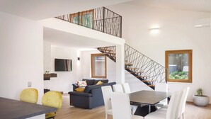 Ein modernes Wohnzimmer mit Essbereich, einer Treppe mit geometrischer Geländerkonstruktion und einem großen Fenster mit Blick auf Grünflächen.