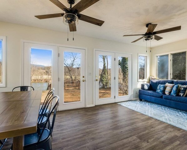 A spacious living area with a wooden dining table, blue sofa, and two ceiling fans. - Serene River Retreat | Double Decks, Hot Tub, Fire Pit & Scenic River Views (Shenandoah)