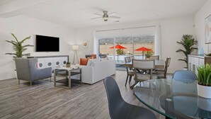 A modern living room with a sofa, a glass coffee table, and a TV mounted on the wall. There is a dining area with a table and chairs, and a large window with red umbrellas outside. - Rolling Hills: Luxury Home, Heated Pool, Mtn Views (Desert Hot Springs)