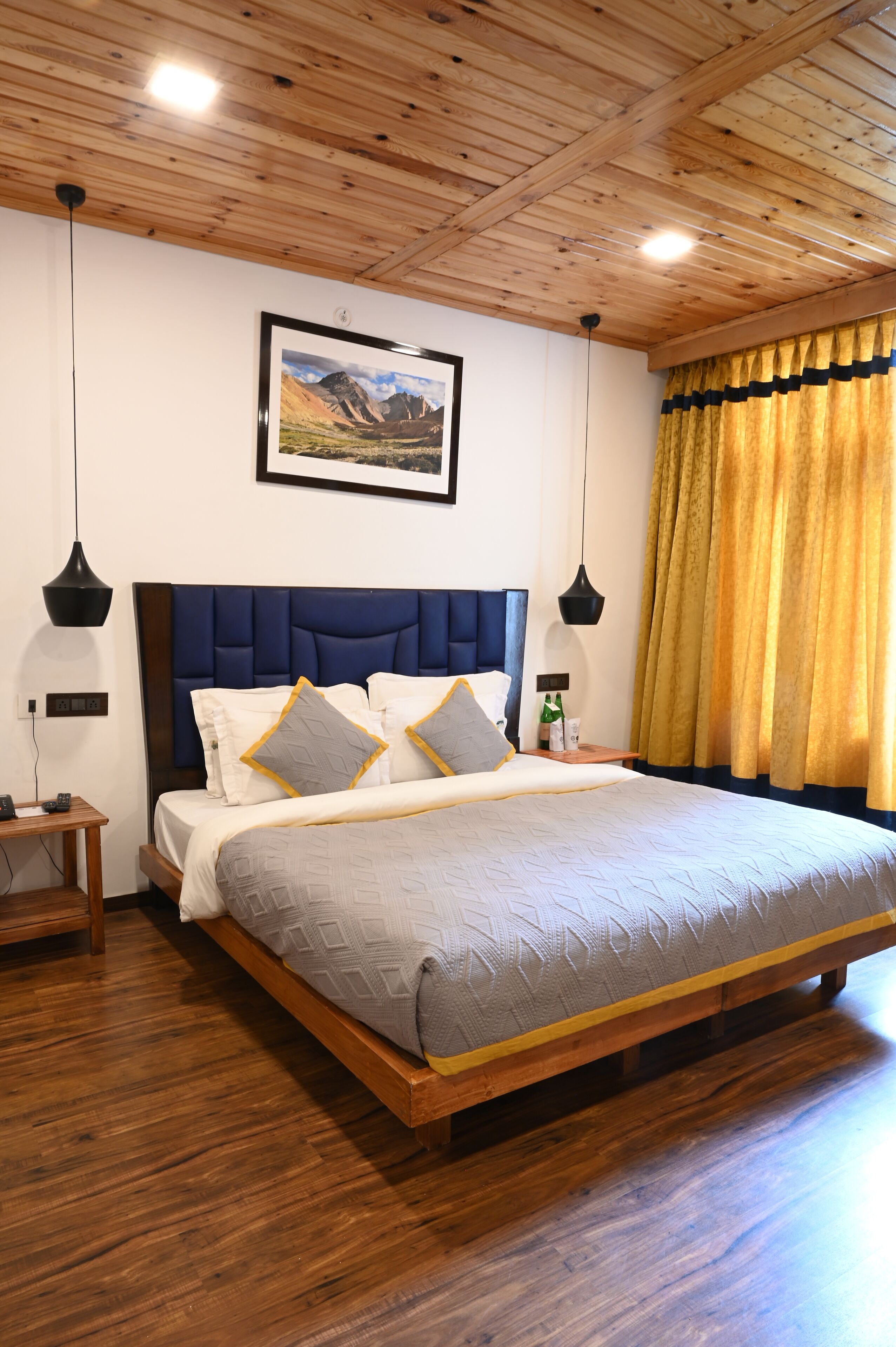 A bedroom with a wooden ceiling, a large bed with a blue headboard, two bedside tables, and a framed picture on the wall.