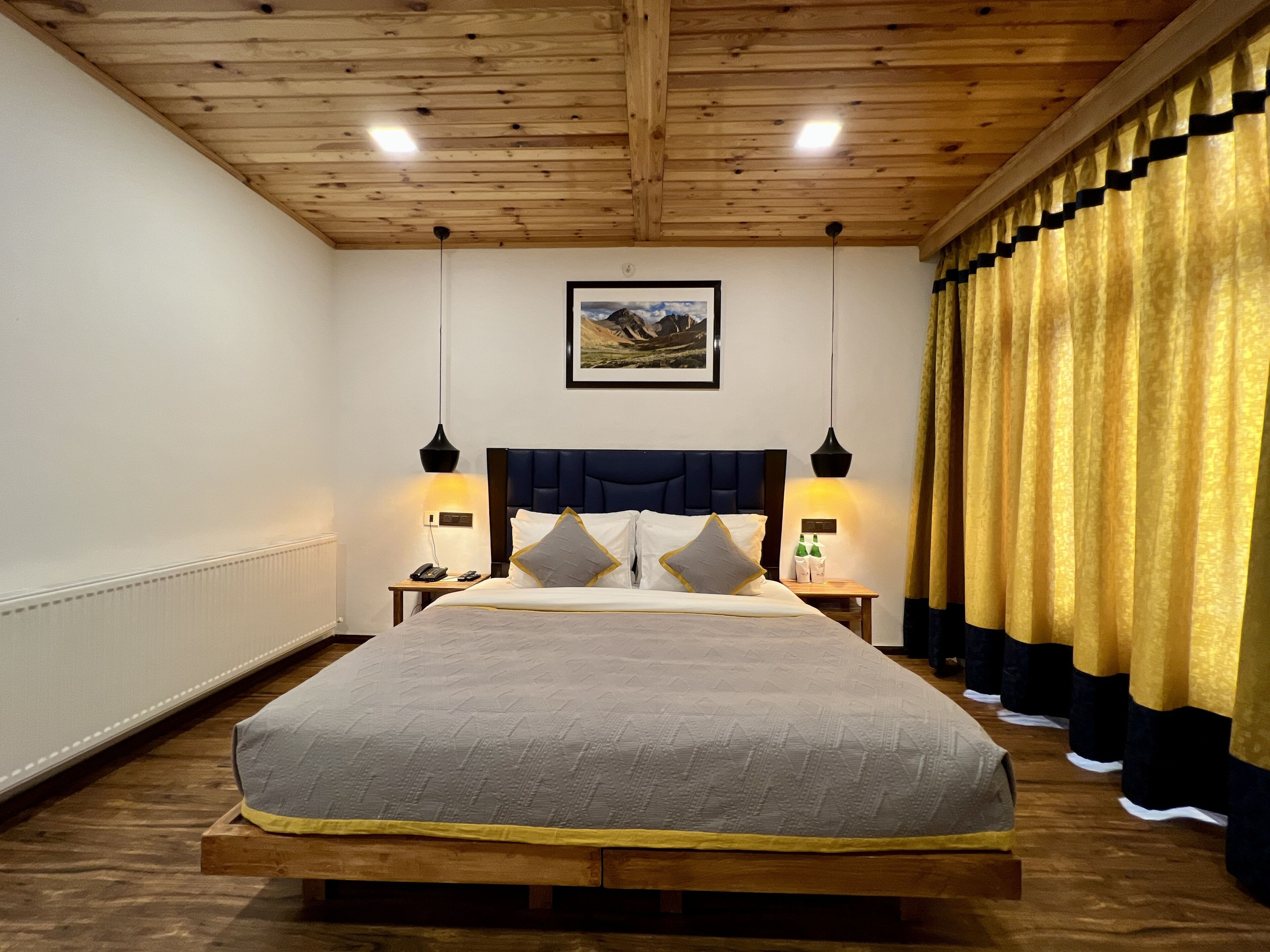 A bedroom with a wooden ceiling, a bed with a dark headboard, two bedside lamps, a framed picture on the wall, and a curtain with a gold and black pattern.