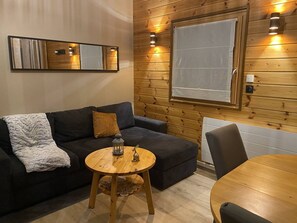Et hyggeligt rum med en mørk sofa, et rundt træbord med en lygte, og et stort vindue med en hvid persienne.
