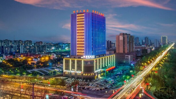 A high-rise hotel with a modern design, featuring a prominent vertical light strip and a large parking area in front. - Ming Hua Hotel (Aksu)