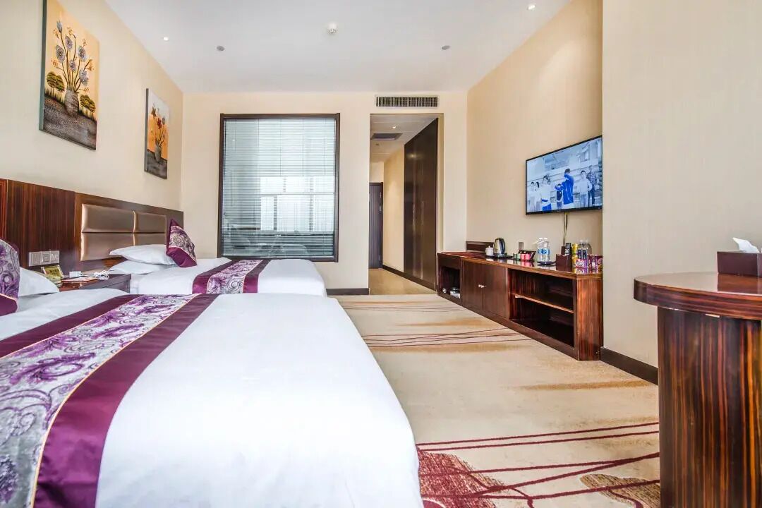 A hotel room with three beds, a TV, a desk, and a large window.