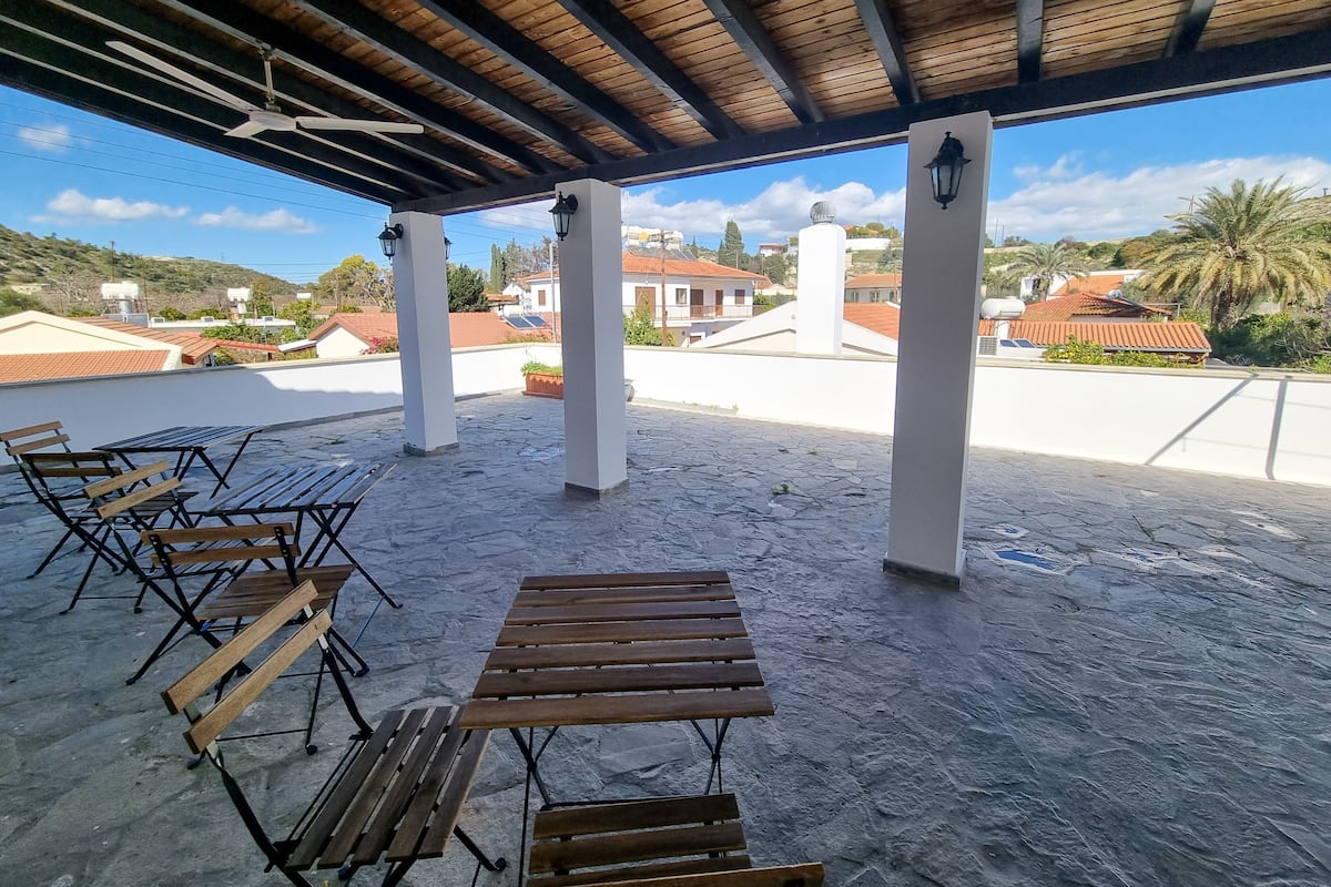 Eine Terrasse im Freien mit Holztischen und -stühlen, einem Deckenventilator und Blick auf umliegende Gebäude.