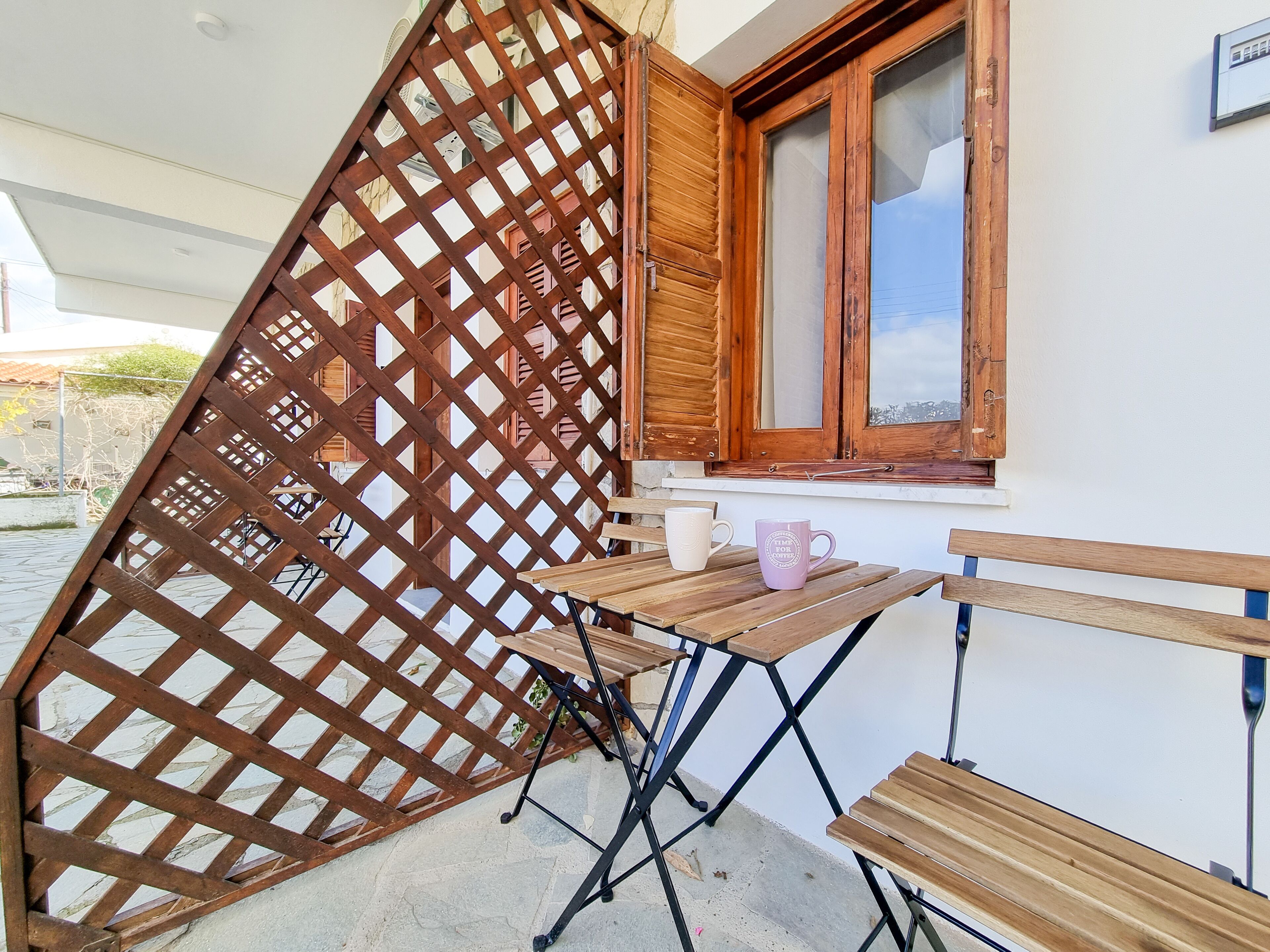 Una mesa de madera con dos tazas, un ventanal con contraventanas de madera y un separador de celosía.