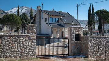 Una villa de paredes de piedra con techo de tejas, una puerta y montañas al fondo.