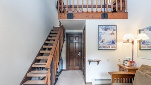 Una escalera con escalones y barandilla de madera, una puerta, una lámpara de pared, un cuadro enmarcado y una mesita con una lámpara.