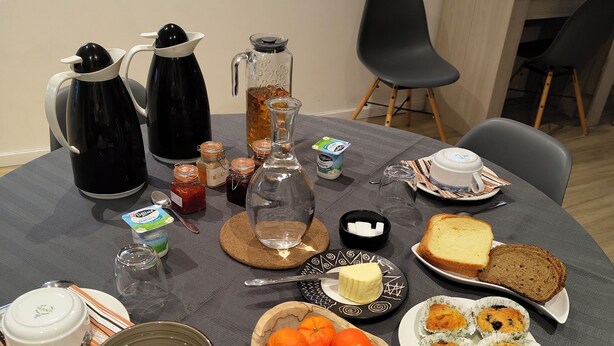 Une table de petit-déjeuner avec des machines à café, un pichet, un bol d’oranges, des muffins, des toasts et une tasse de thé.
